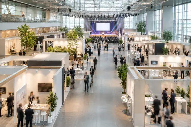 Belebte Messehalle mit weißen Ausstellungsständen, Besuchern im Gespräch und einer beleuchteten Konferenzbühne im Hintergrund.