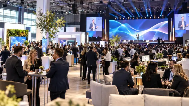 Messehalle mit Glasfront, in der ein Mann auf einer beleuchteten Bühne vor einer Leinwand mit blauen Wellenlinien spricht, während sich im Vordergrund Besucherinnen und Besucher an Stehtischen und auf Sofas zwischen Ausstellungsständen und Pflanzen unterhalten.